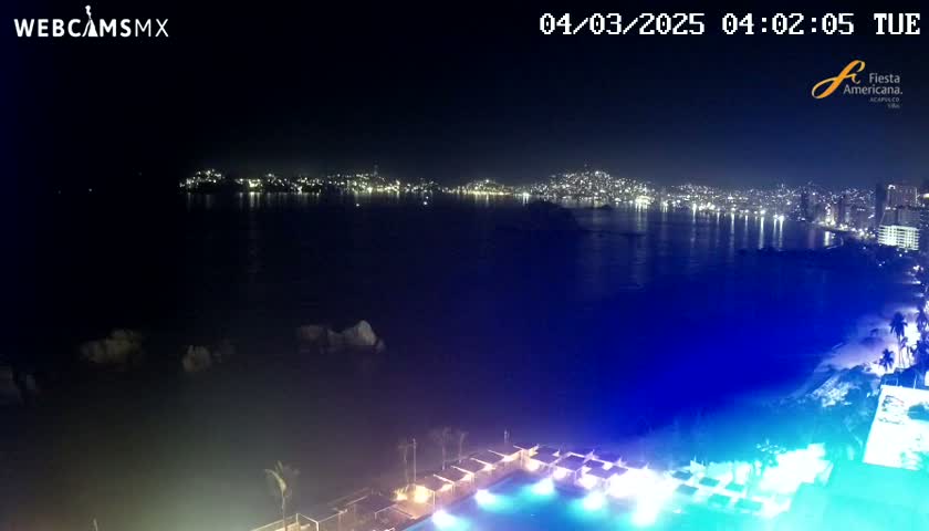 A nighttime view of a city's illuminated coastline across a dark body of water, with a brightly lit pool and cabanas in the foreground.