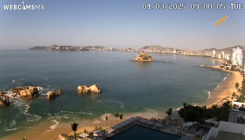 A sunny coastal scene shows a city across a bay, a sandy beach with gentle waves, and rocky outcrops in the foreground, all under a clear sky.