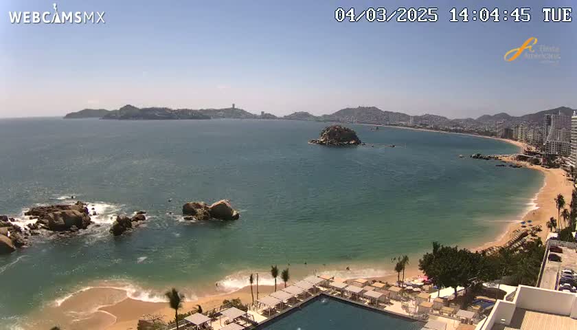 A sunny day reveals a coastline with a sandy beach, calm ocean waters, rocky outcrops, and a resort with a swimming pool overlooking the scene.