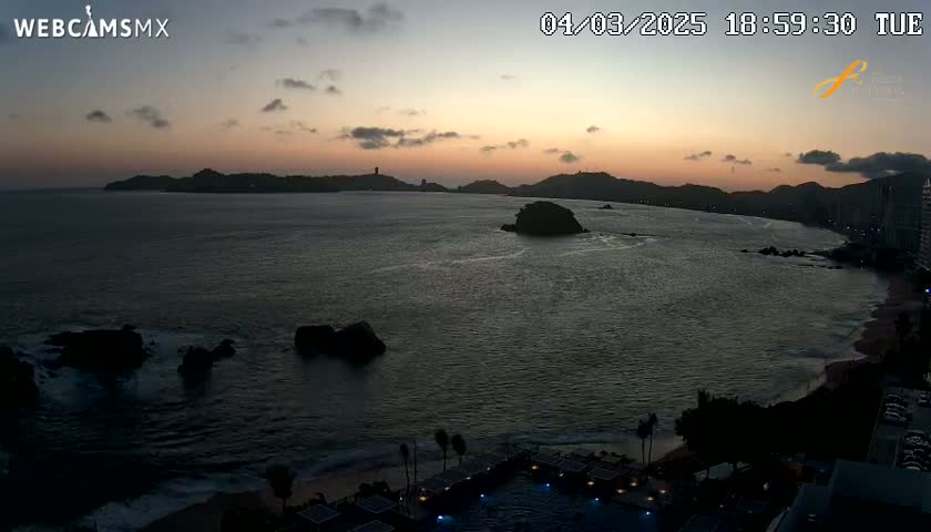 An ocean sunset view shows dark silhouettes of coastal mountains and rocks along a beach at dusk.