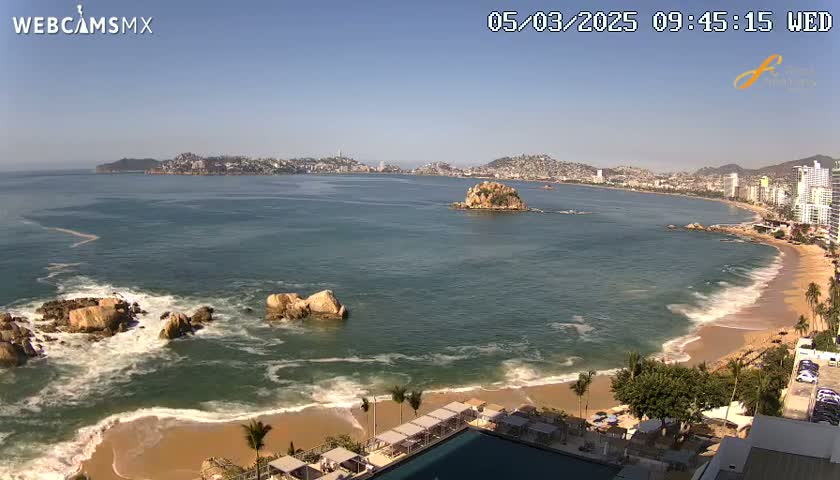 A sunny coastal scene shows a calm ocean with waves breaking on a sandy beach, rocky outcrops, and a city in the distance.