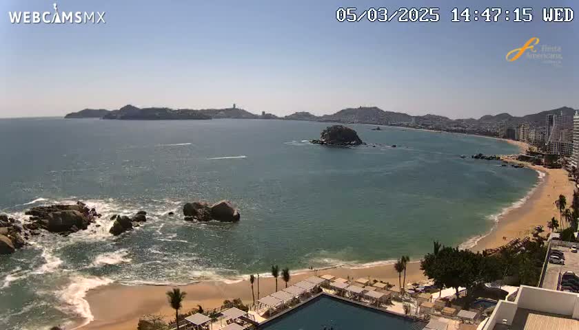 A sunny day shows a beach with calm waves lapping a sandy shore, with rocky outcrops in the ocean and a city skyline in the distance.