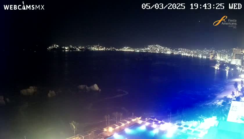 A nighttime view of a city skyline across a dark body of water, with a pool and illuminated cabanas in the foreground.