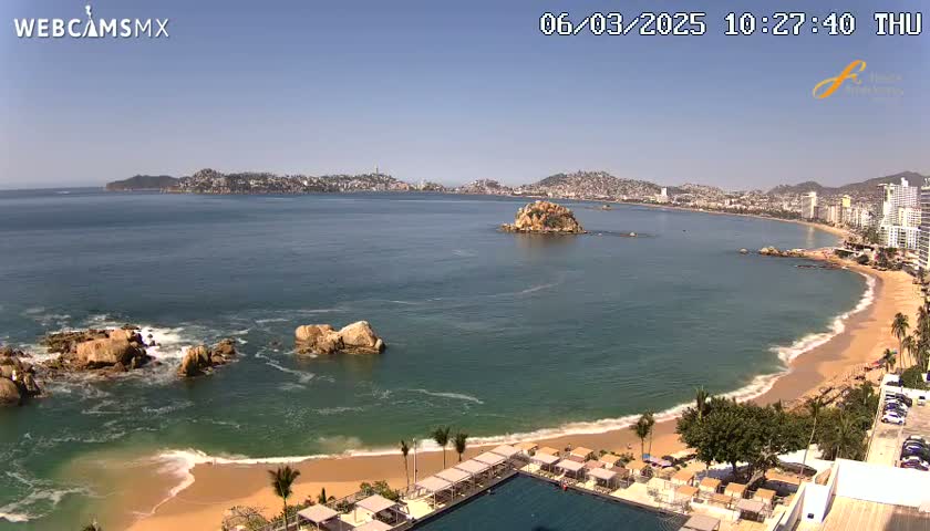 A sunny day at a beach with calm, clear water, showing a sandy shore, rocky outcrops, and a distant city skyline.