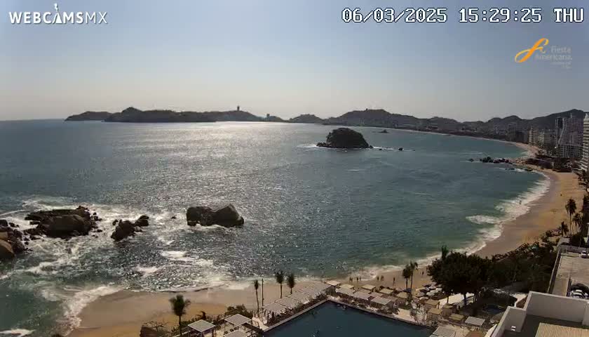A sunny day reveals a beach with calm waves lapping the shore, rocky outcrops in the water, and a coastal city visible in the distance.