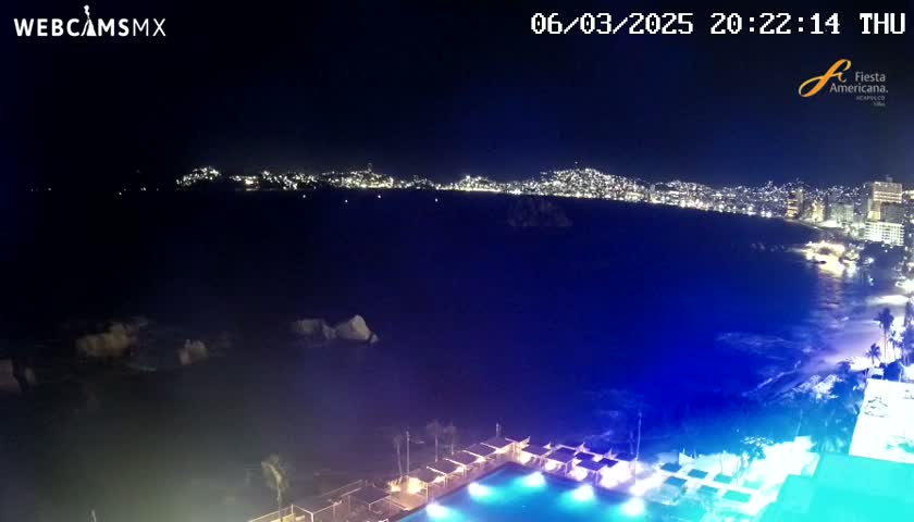 A nighttime view of a city's illuminated coastline from an elevated vantage point, overlooking a resort pool area with the ocean in the foreground.