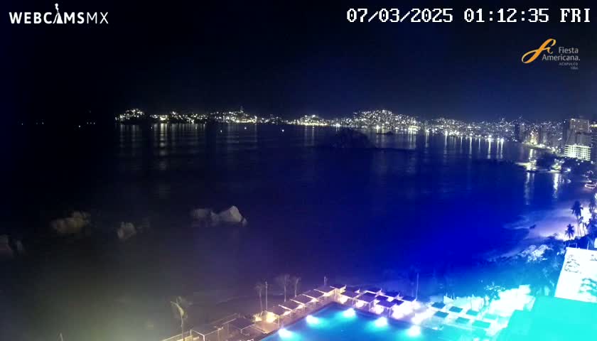A nighttime view of a city skyline across a dark-blue ocean, with a brightly lit pool and beach in the foreground.