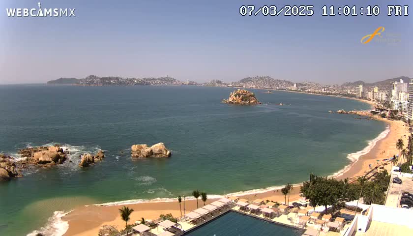 A sunny beach with calm, teal water curves around a rocky point, showcasing a city skyline in the distance.