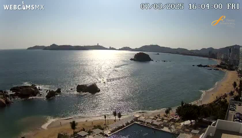 A sunny day at a beach with calm, sparkling water, rocky outcrops, a sandy shore, and buildings along the coastline.