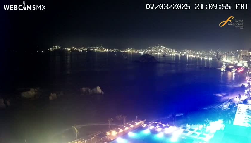 A nighttime view of a city's lights across a dark body of water, with a brightly lit pool and beach area in the foreground.