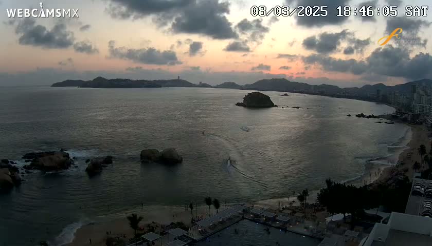 An expansive ocean bay at sunset, with a beach, buildings, and several small islands visible under a partly cloudy sky.