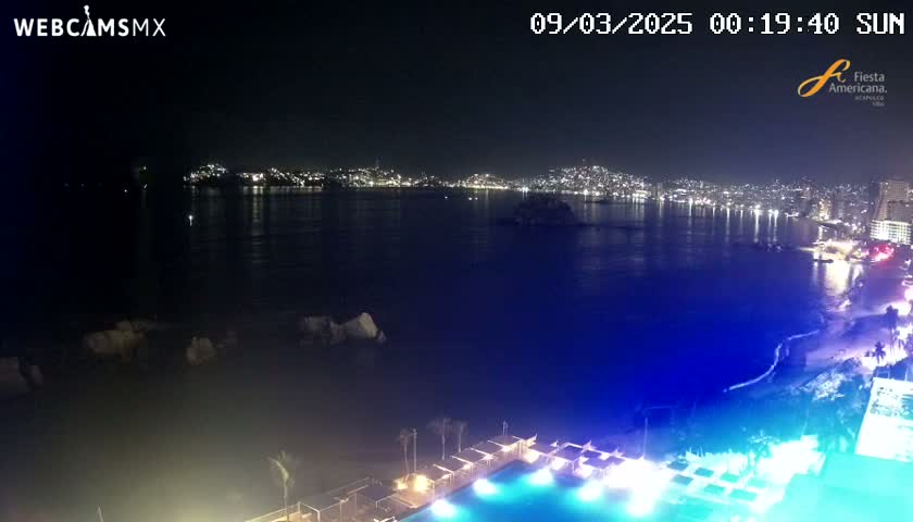 A nighttime view of a city across a dark body of water, with a brightly lit pool and beach in the foreground under a clear sky.