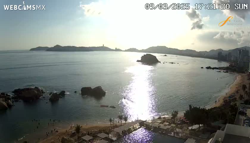 A partly sunny afternoon scene shows a calm ocean bay with rocky outcrops, a sandy beach, and a resort with a swimming pool, all set against a backdrop of distant hills.