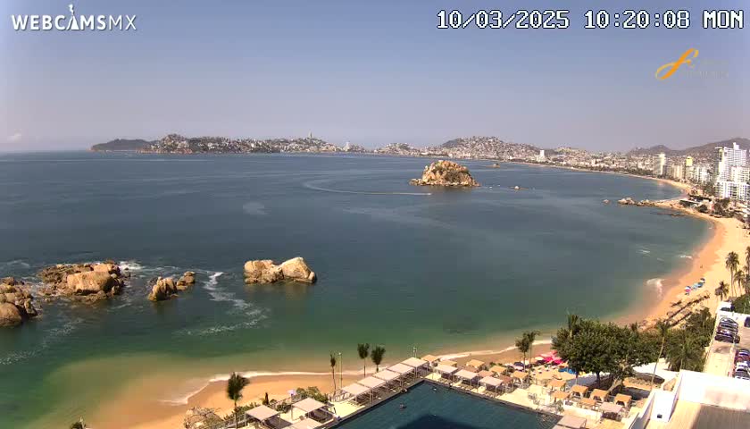 A sunny day overlooking a calm bay with a sandy beach, rocky outcrops, and a distant city skyline.