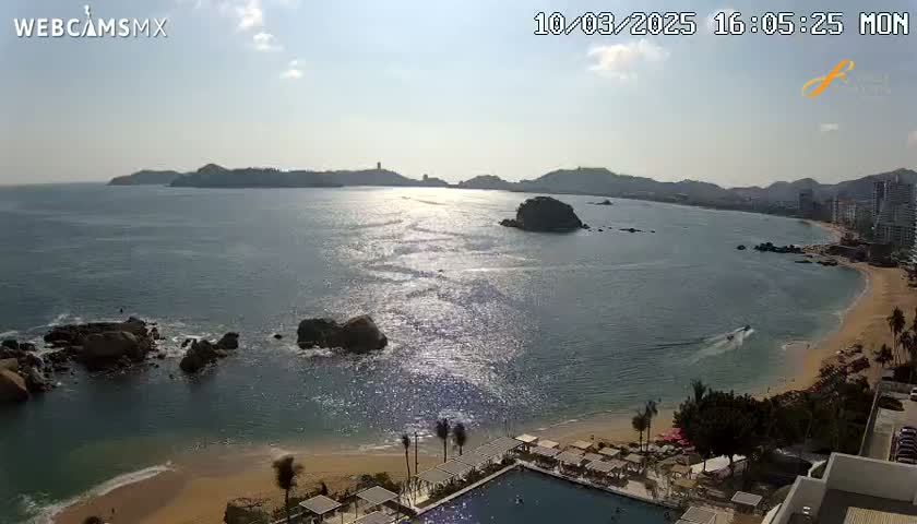 A sunny day reveals a coastal scene with a calm ocean reflecting sunlight, a sandy beach, rocky outcroppings, and distant hills under a mostly clear sky.