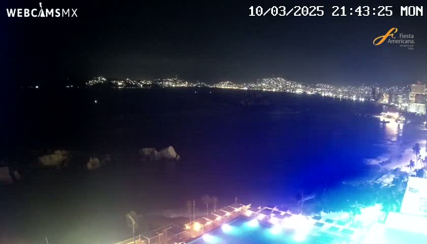 A nighttime view of a city skyline across a dark body of water, with a brightly lit pool and beach area in the foreground.