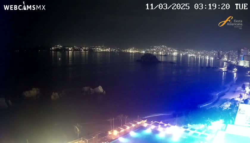 A nighttime coastal view shows a city's lights reflected on a calm body of water, with a brightly lit pool and beach in the foreground.