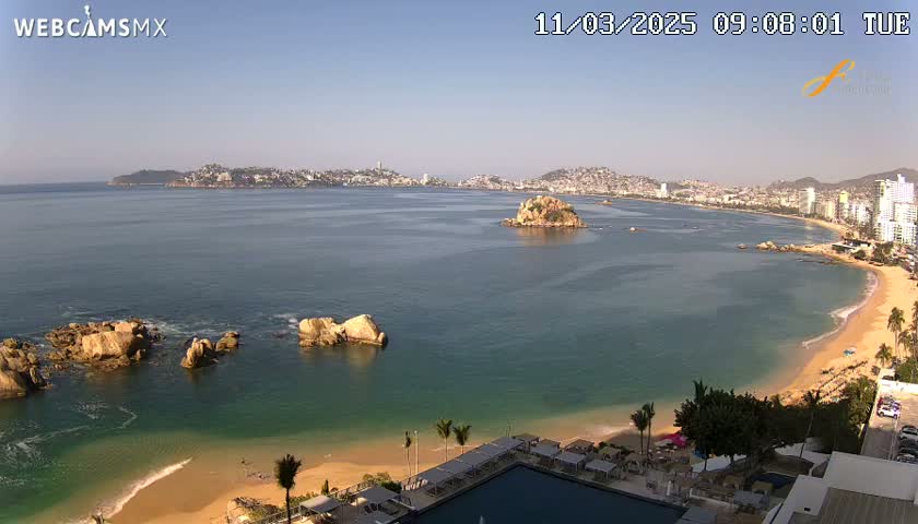 A sunny coastal scene shows a sandy beach curving around a calm bay with rocky outcrops, a small island, and a city in the distance.