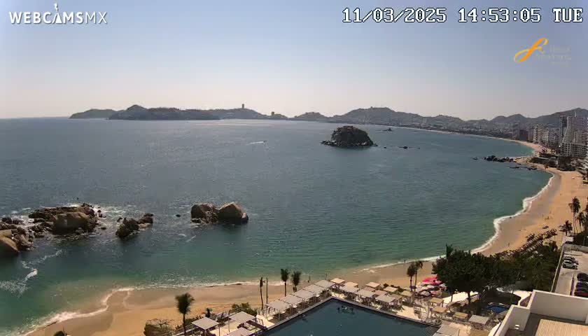 A sunny, coastal scene shows a calm ocean bay with a sandy beach, rocky outcrops, and distant hills under a clear sky.