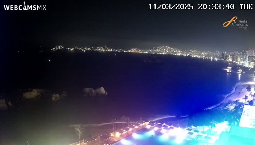 A nighttime coastal view shows a city skyline across a dark body of water, with illuminated beachside structures in the foreground under a clear sky.