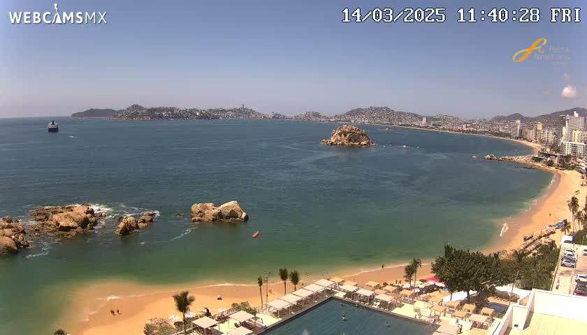 An aerial view shows a sunny beach with calm, teal water, rocky outcrops, a resort with a swimming pool, and a distant city skyline.