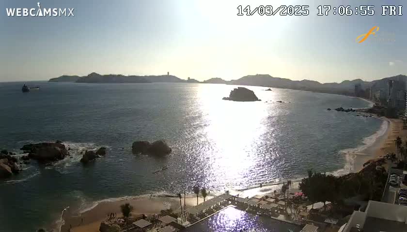 A sunny, high-angle view shows a calm ocean bay with a sandy beach, rocky outcroppings, distant mountains, and a resort with a swimming pool.