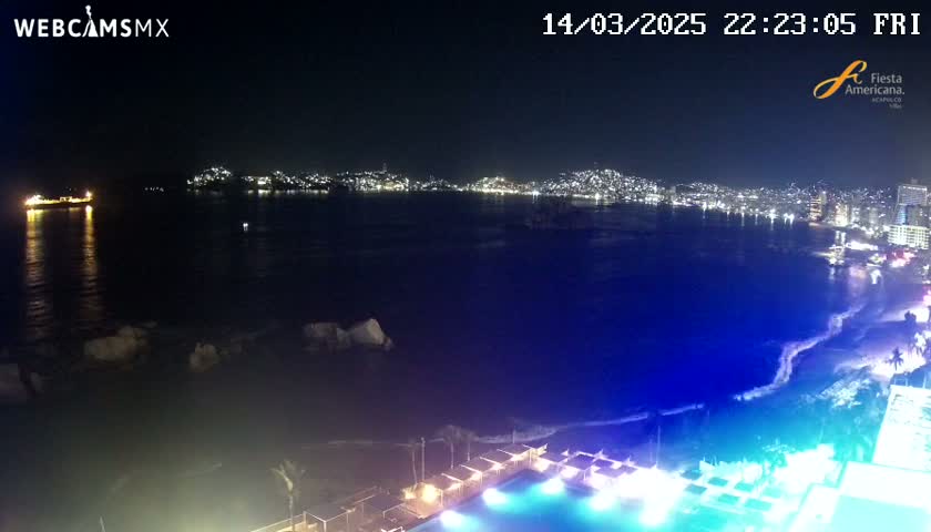A nighttime view of a city skyline across a dark bay, with a brightly lit pool and beach in the foreground and gentle waves.
