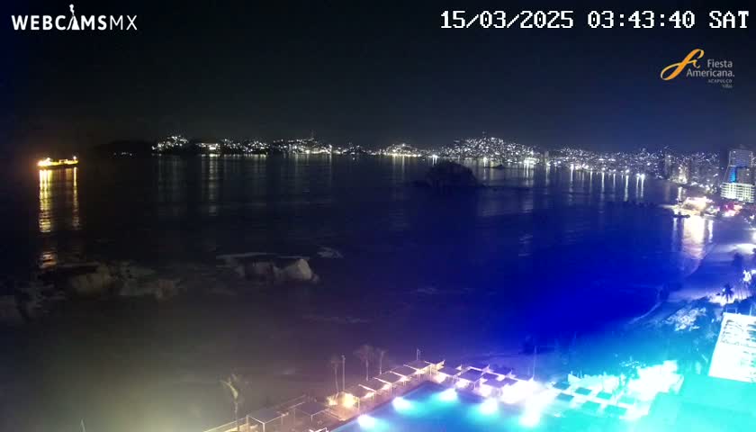 A nighttime view of a city skyline across a dark, calm body of water, with a brightly lit pool and cabanas in the foreground.