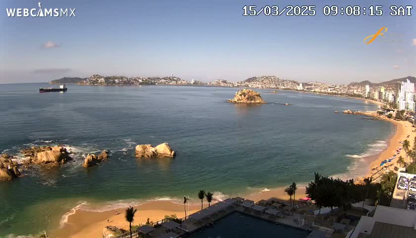 A sandy beach with calm, clear water and a distant city skyline under a clear blue sky is visible from a high vantage point.