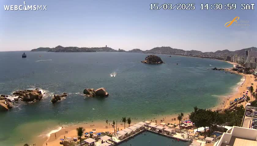A sunny, high-angle view shows a calm bay with a sandy beach, rocky outcrops, and buildings along the shore.