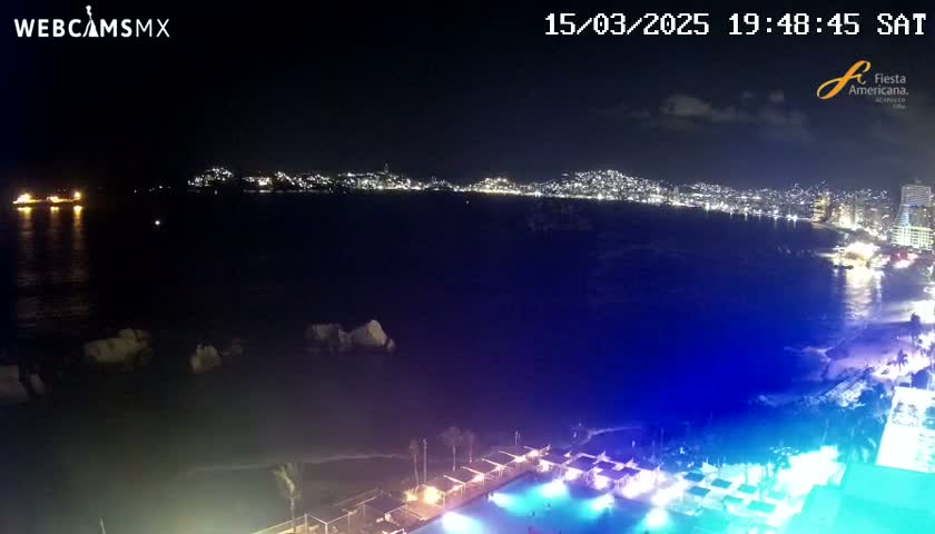 A nighttime view of a city's illuminated coastline across a dark ocean, with a brightly lit pool and beach area in the foreground.