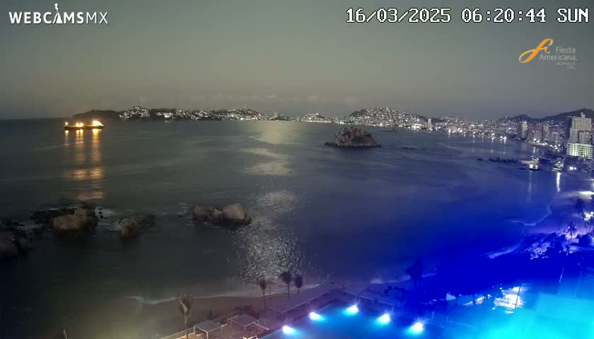A nighttime coastal scene shows a dark body of water with a city skyline in the distance, a ship with lights on, some rocks near the shore, and a pool lit in blue at the bottom.