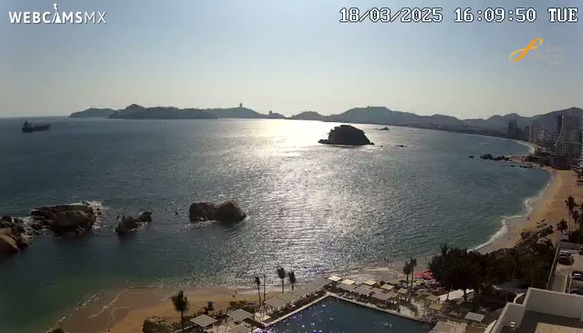 A sunny, high-angle view of a calm bay with a sandy beach, rocky outcrops, distant hills, and a resort with a swimming pool.