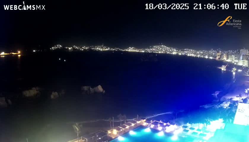 A nighttime coastal view shows a city skyline across a dark body of water, with a brightly lit pool and surrounding structures in the foreground.