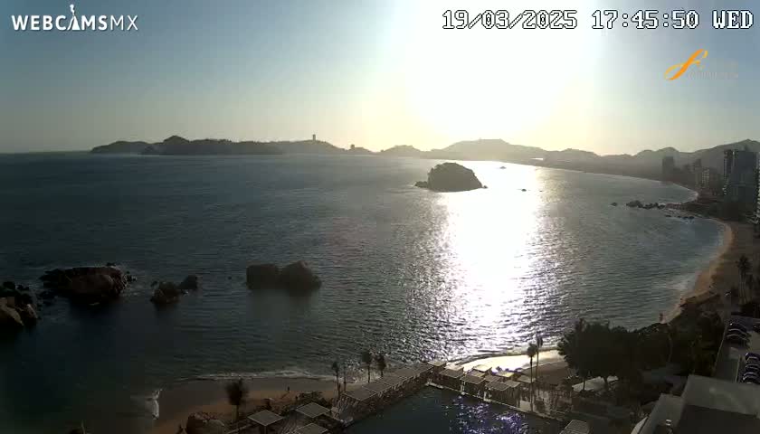 A tranquil ocean scene at sunset, showing a sandy beach, rocky outcrops, and distant hills under a clear sky.
