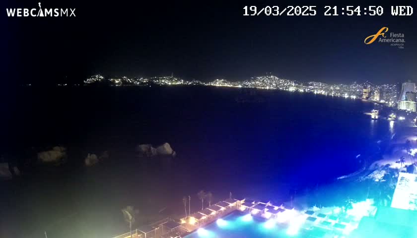 A nighttime view of a city skyline across a dark body of water, with a brightly lit pool and surrounding structures in the foreground under a clear sky.