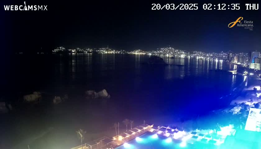 A nighttime view of a city's illuminated coastline across a dark body of water, featuring a poolside area with illuminated cabanas in the foreground.