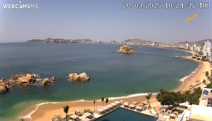 A beach with calm, clear water and a resort is shown under sunny skies, with a city and more coastline visible in the distance.