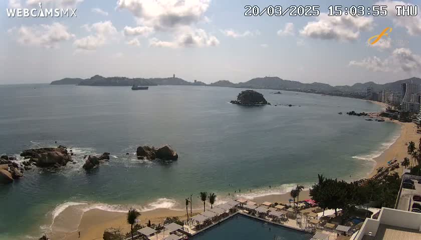 A partly cloudy day shows a view of a bay with a sandy beach, rocky outcrops, and distant hills, all under a mostly sunny sky.