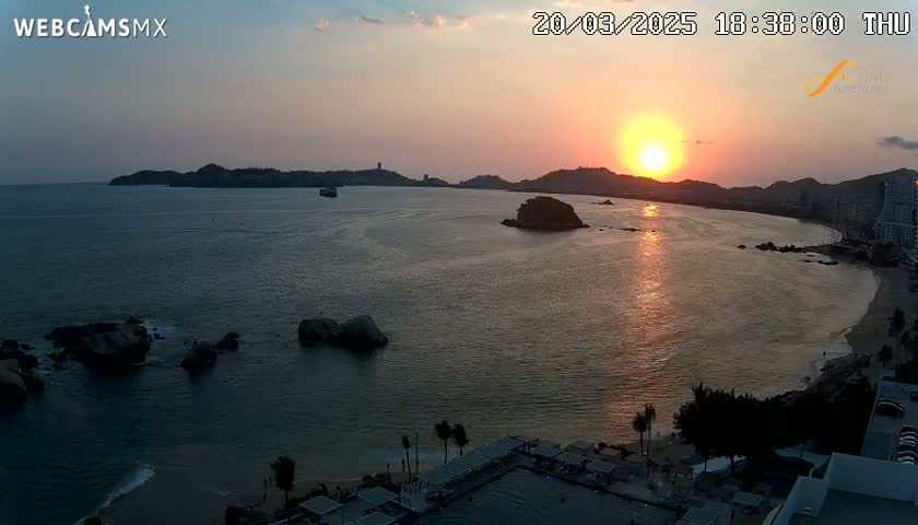 A sunset over a calm ocean with a beach and rocky outcrops, silhouetted hills in the background.