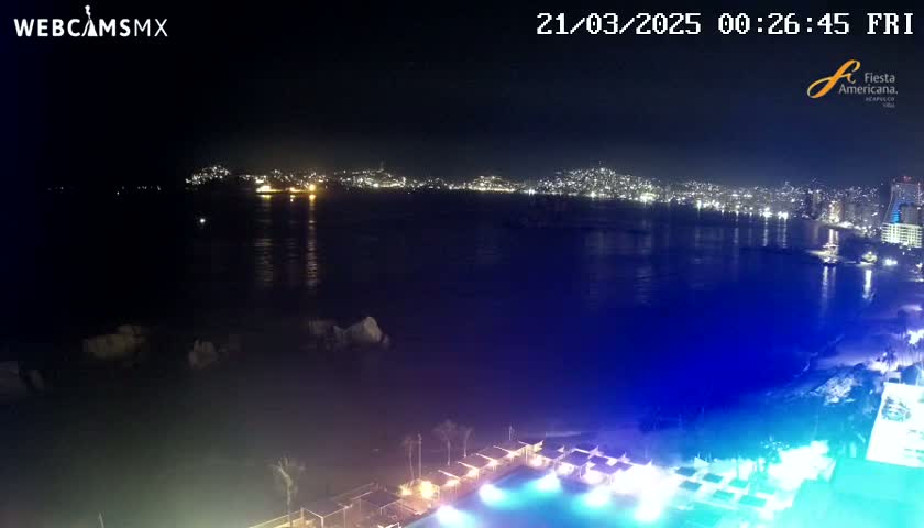 A nighttime view of a city skyline across a calm body of water, with a brightly lit pool and beach area in the foreground.