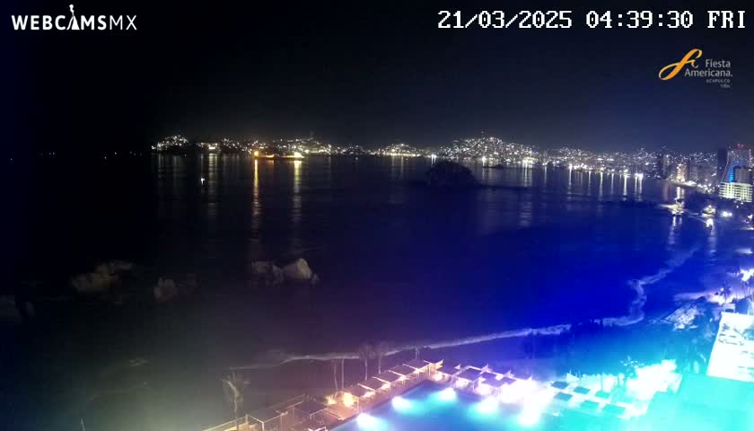 A nighttime view of a city skyline across a dark, calm ocean, with a brightly lit pool and beach in the foreground.