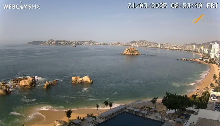 A sunny coastal scene shows a sandy beach, calm ocean water, rocky outcrops, and a distant city skyline.