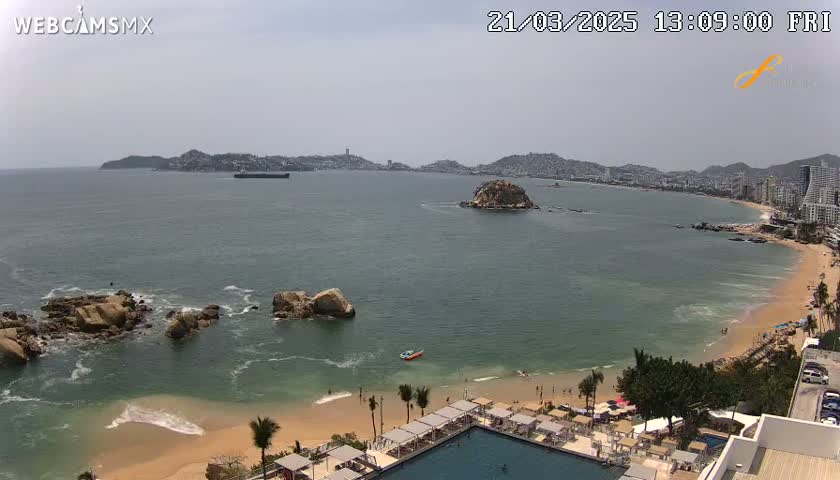 A beach with calm ocean waters, under a partly cloudy sky, is viewed from an elevated position showing a resort area and distant city.