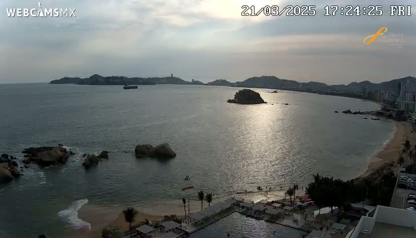 A tranquil coastal scene under a partly cloudy sky shows a calm bay with a sandy beach, rocky outcrops, and buildings along the shoreline at sunset.