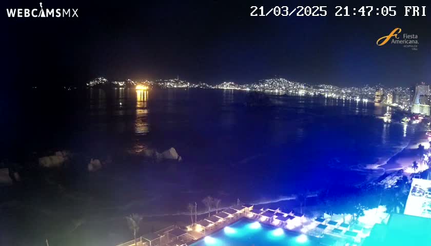 A nighttime view of a city skyline across a dark, wavy ocean, with a beach and illuminated pool in the foreground.