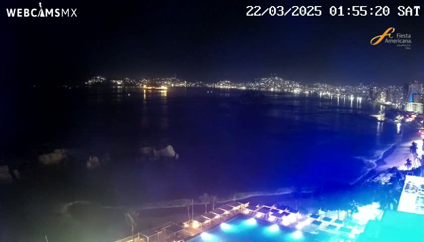 A nighttime view of a city skyline across a dark-blue ocean, with a brightly lit pool and beach area in the foreground.