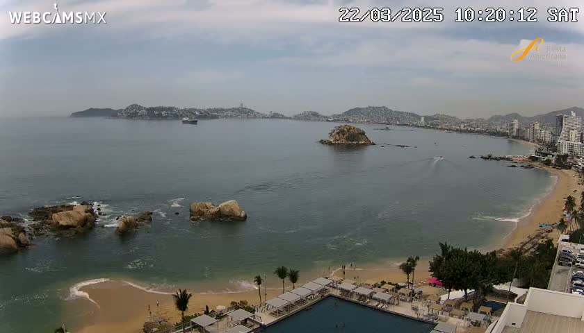 An overcast day reveals a coastal scene with a sandy beach, calm ocean waters, rocky outcrops, a resort with a pool, and a distant cityscape under a cloudy sky.