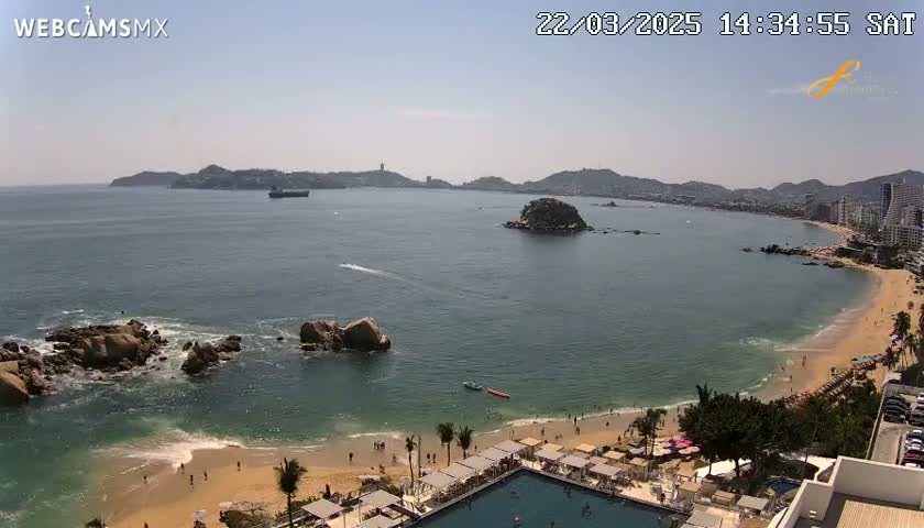 A sunny, expansive view of a bay with a sandy beach, rocky outcrops, and distant hills, showing a resort area with a swimming pool and people enjoying the beach.