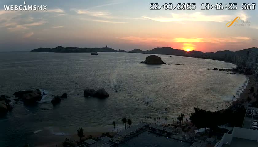 A sunset over a calm bay with rocky outcrops, a beach, and buildings along the shoreline.